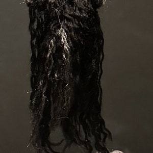 Puede incluir: Una mu&ntilde;eca de crochet con el pelo largo negro y un vestido de crochet gris. La mu&ntilde;eca est&aacute; de pie sobre una caja de crochet marr&oacute;n y gris.