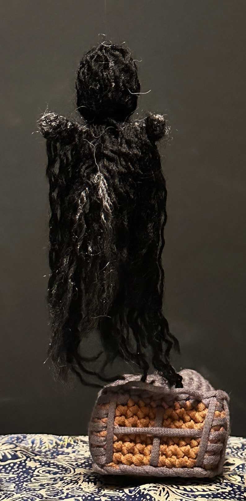 Puede incluir: Una mu&ntilde;eca de crochet con el pelo largo negro y un vestido de crochet gris. La mu&ntilde;eca est&aacute; de pie sobre una caja de crochet marr&oacute;n y gris.