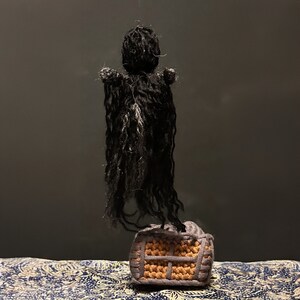 Puede incluir: Una figura negra de crochet con una larga capa fluida se encuentra sobre una casa de crochet marr&oacute;n y naranja. La figura est&aacute; suspendida del techo.
