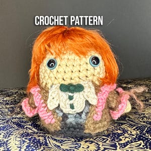 May include: Crocheted amigurumi doll with orange hair, brown and pink clothing, and blue eyes. The doll is sitting on a patterned fabric.