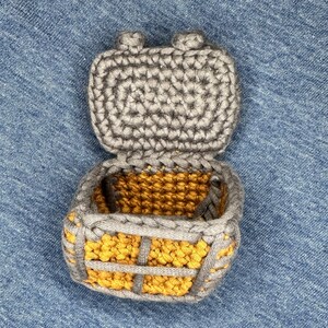 Puede incluir: Una caja de crochet con una tapa gris y un interior amarillo. La tapa est&aacute; abierta y la caja est&aacute; sobre un fondo de mezclilla azul.