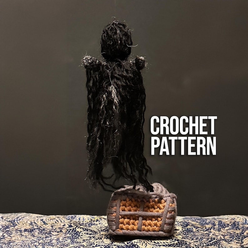 Puede incluir: Patr&oacute;n de crochet para una figura negra con capucha de pie sobre una peque&ntilde;a casa de crochet. La figura tiene el pelo largo y suelto y una capa negra. La casa es marr&oacute;n y tiene una peque&ntilde;a ventana.