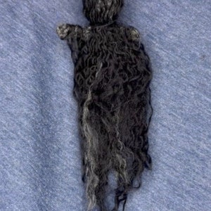 Puede incluir: Una mu&ntilde;eca de lana negra con el pelo largo y en forma de cuerda. La mu&ntilde;eca est&aacute; hecha de lana y tiene un dise&ntilde;o simple y minimalista.