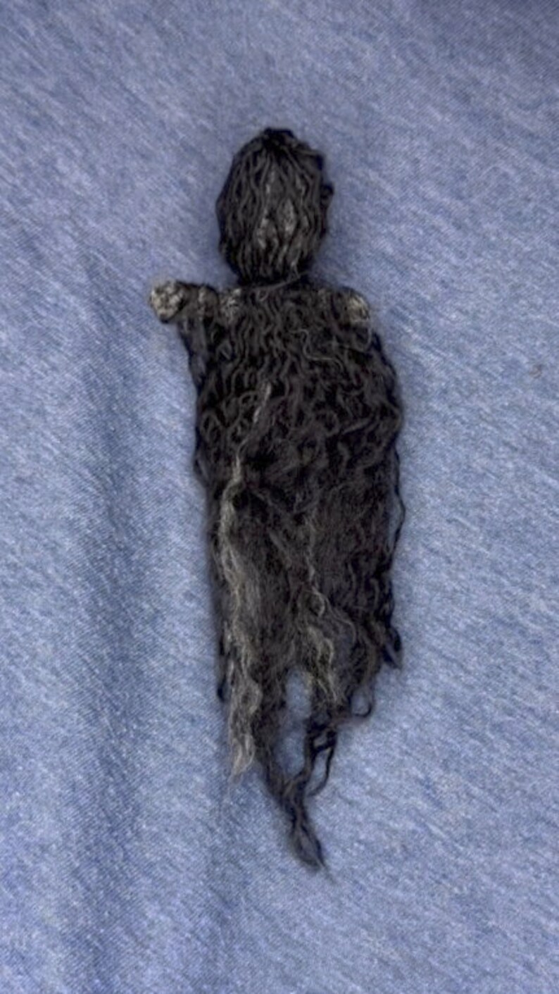 Puede incluir: Una mu&ntilde;eca de lana negra con el pelo largo y en forma de cuerda. La mu&ntilde;eca est&aacute; hecha de lana y tiene un dise&ntilde;o simple y minimalista.