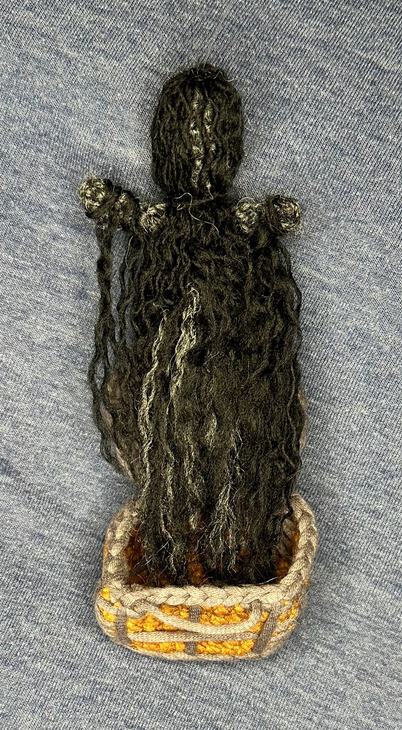 Puede incluir: Una mu&ntilde;eca de crochet con el pelo largo y negro, que lleva una cesta de crochet gris y amarilla.