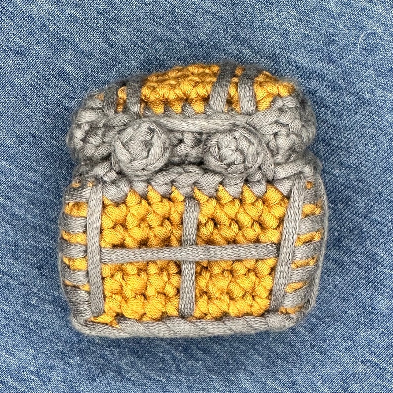 Puede incluir: Un adorno de casa tejido a crochet en color amarillo y gris. La casa tiene una peque&ntilde;a ventana y un techo con dos detalles redondeados.