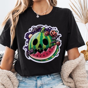 May include: Black t-shirt with a graphic of a watermelon with a jack-o'-lantern face and bats flying around it. The watermelon is green and red with a black outline.