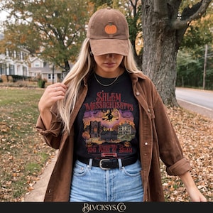 May include: A brown corduroy baseball cap with an embroidered orange pumpkin, worn with a black t-shirt featuring a graphic of Salem, Massachusetts, a witch on a broomstick, and the text Celebrating 400 Years of Salem, paired with a brown corduroy shirt.