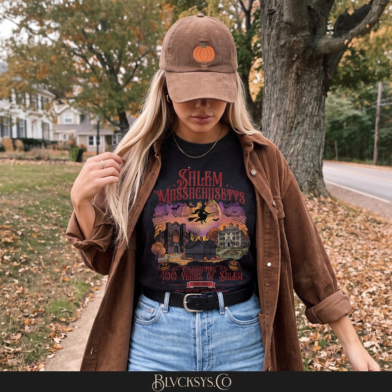 May include: A brown corduroy baseball cap with an embroidered orange pumpkin, worn with a black t-shirt featuring a graphic of Salem, Massachusetts, a witch on a broomstick, and the text Celebrating 400 Years of Salem, paired with a brown corduroy shirt.