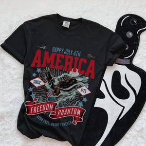 May include: Black t-shirt with a red, white and blue graphic design featuring an eagle, stars, and the text "Happy July 4th America Since 1776 Freedom Phantom Born Free, Haunt Forever".