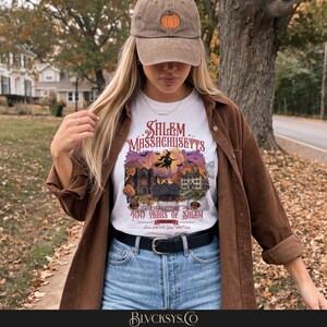 May include: A white t-shirt featuring a graphic of Salem, Massachusetts, with a witch on a broomstick, historic buildings, and the text Celebrating 400 Years of Salem. The shirt is worn with a brown corduroy shirt and a tan baseball cap with a pumpkin.