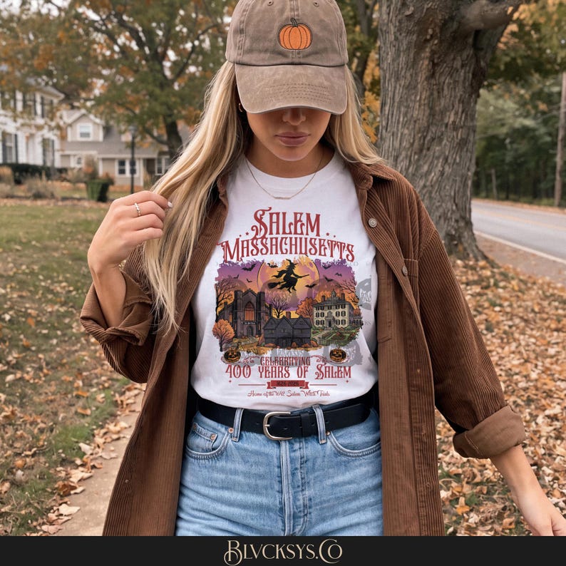 May include: A white t-shirt featuring a graphic of Salem, Massachusetts, with a witch on a broomstick, historic buildings, and the text Celebrating 400 Years of Salem. The shirt is worn with a brown corduroy shirt and a tan baseball cap with a pumpkin.