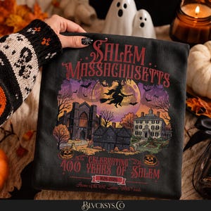 May include: A black t-shirt featuring a graphic of Salem, Massachusetts, with a witch on a broomstick flying in front of a large moon. The design includes historic buildings, pumpkins, and text reading Celebrating 400 Years of Salem 1626-2026.