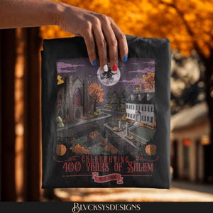 May include: A black t-shirt featuring a graphic illustration of a town scene with a large stone church, a white colonial house, pumpkins, and a witch flying across a full moon. The text reads Celebrating 400 Years of Salem 1626-2026.