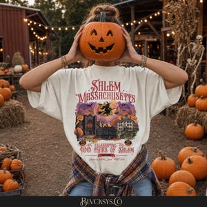 May include: An off-white t-shirt featuring a graphic of Salem, Massachusetts, with a witch on a broomstick, historic buildings, and the text Celebrating 400 Years of Salem 1626-2026. The shirt is worn with a plaid flannel shirt tied around the waist.