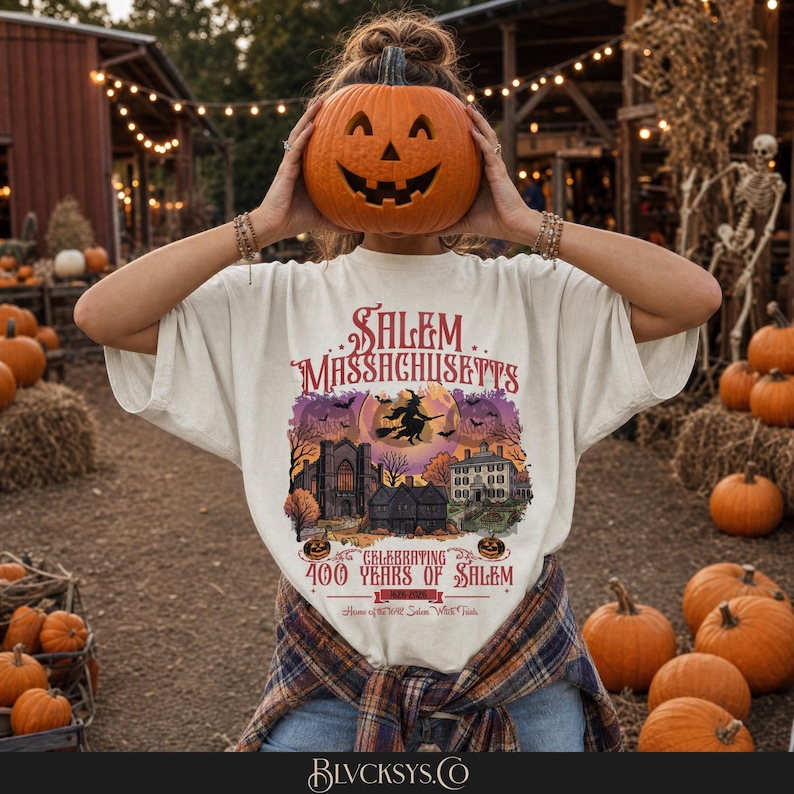May include: An off-white t-shirt featuring a graphic of Salem, Massachusetts, with a witch on a broomstick, historic buildings, and the text Celebrating 400 Years of Salem 1626-2026. The shirt is worn with a plaid flannel shirt tied around the waist.