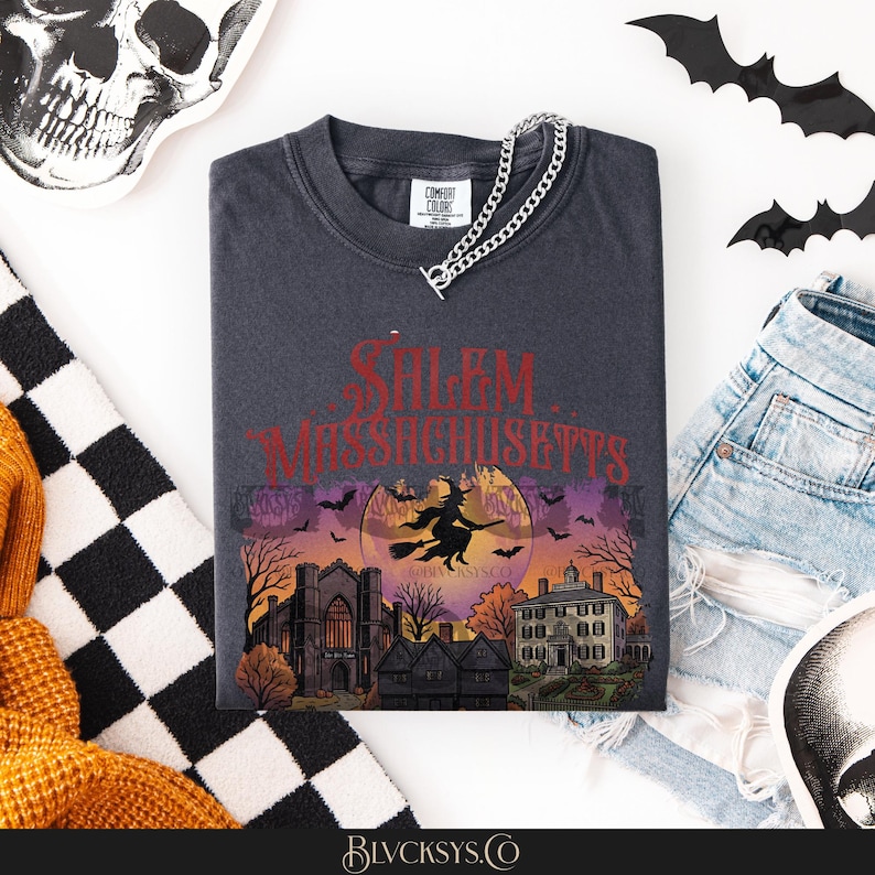 May include: A charcoal gray t-shirt featuring a graphic print of a witch flying on a broomstick in front of a large moon, with the text Salem Massachusetts above. The design includes silhouettes of buildings, trees, and bats in a sunset color palette.