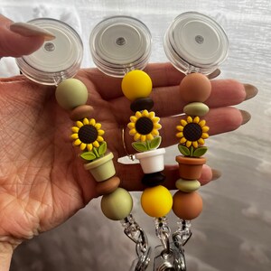 Könnte beinhalten: Drei einziehbare Ausweishalter mit Sonnenblumen- und Topf-Anhängern. Jeder Ausweishalter hat ein transparentes Kunststoffgehäuse, einen Metallclip und eine Perlenkette mit braunen, grünen und gelben Perlen.