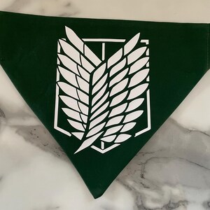May include: Dark green bandana with a white emblem featuring a stylized wing design inside a geometric frame. The bandana is triangular in shape and made of a smooth fabric. The emblem is centered on the bandana.