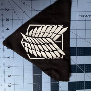 May include: A black triangular bandana with a white winged symbol, resembling a stylized bird, in the center. The symbol is surrounded by a white square. The bandana is on a blue gridded surface.