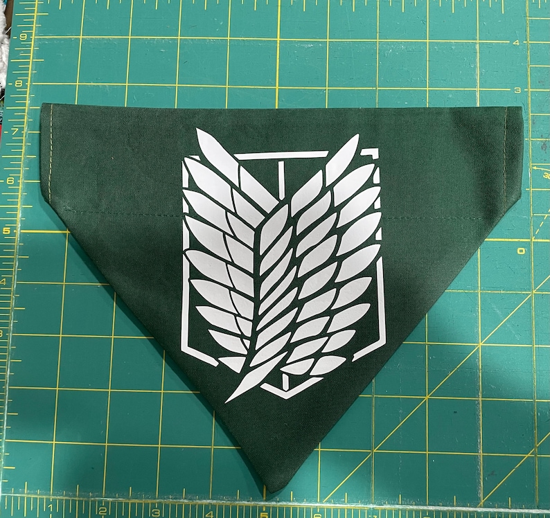 May include: A dark green bandana with a white emblem featuring a stylized wing design. The bandana is triangular in shape and made of fabric. The emblem is centered on the bandana.