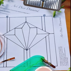 May include: A stained glass pattern for an Art Deco Diamond design. The pattern is printed on white paper and includes a black diamond shape with lines radiating from the center. A green glass cutter is lying on the paper. The text "Art Deco Diamond" is written on the paper.