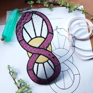 Op de afbeelding: Een zonneslangontwerp van glas-in-lood met een paars en geel kleurenschema. Het stuk hangt aan een zwart touwtje en is omgeven door een wit vel papier met een zwart-witte omtrek van het ontwerp. De tekst "Sol Serpent" staat op het papier.