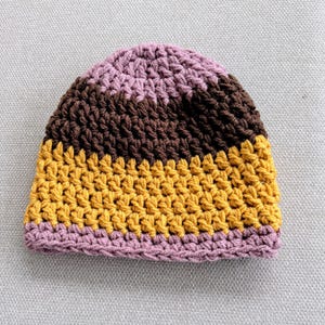 May include: A crochet beanie hat featuring horizontal stripes in alternating colors of light purple, dark brown, and mustard yellow. The hat is constructed with a thick, textured yarn stitch pattern, creating a rounded shape with a ribbed edge.