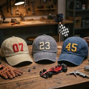 May include: Three baseball caps with embroidered numbers: 07 in red, 23 in white, and 45 in yellow. The caps are in tan, gray, and blue. A miniature race car, a checkered flag, and a pair of brown gloves are on a wooden surface.