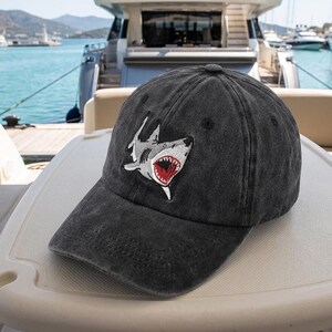 May include: A charcoal gray baseball cap featuring an embroidered patch of a shark with an open mouth, sharp teeth, and a red interior. The cap has a distressed, washed-out fabric texture, a curved brim, and a button at the top center of the crown.