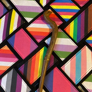 Peut inclure: Une baguette en bois avec une poignée brune et noueuse se trouve sur un patchwork de carrés de tissu. Chaque carré est d'une couleur et d'un motif différents, notamment des rayures arc-en-ciel, roses, violettes et bleues. Les carrés sont cousus ensemble en forme de losange.