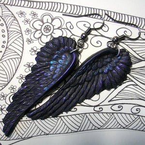 May include: A pair of dark purple wing-shaped earrings with iridescent blue and purple highlights. The earrings are suspended from silver-colored ear wires. The wings have a textured surface and are set against a black and white patterned background.