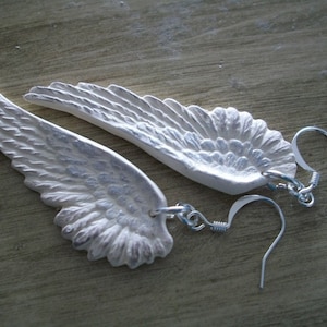 K&ouml;nnte beinhalten: Ein Paar silberne Engelsfl&uuml;gel-Ohrringe mit einem detaillierten Design. Die Fl&uuml;gel sind aus einem strukturierten Material gefertigt und haben eine leicht irisierende Oberfl&auml;che. Die Ohrringe sind an silbernen Haken befestigt.