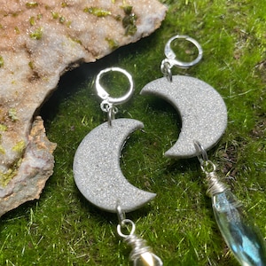 May include: Silver-toned crescent moon earrings with a glittery, textured surface. Each earring features a silver leverback clasp and a dangling, clear, teardrop-shaped charm. The earrings are displayed on a bed of green moss.