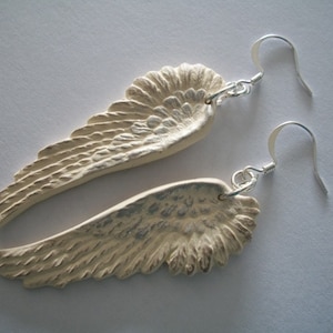 Puede incluir: Un par de pendientes de plata con alas de &aacute;ngel blancas. Las alas son texturizadas y tienen un aspecto ligeramente desgastado.