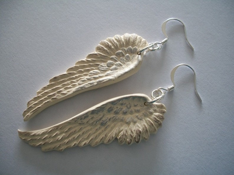 Puede incluir: Un par de pendientes de plata con alas de &aacute;ngel blancas. Las alas son texturizadas y tienen un aspecto ligeramente desgastado.