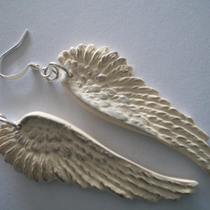 Puede incluir: Un par de pendientes de aro de plata con alas de &aacute;ngel de cer&aacute;mica blanca. Las alas tienen un dise&ntilde;o texturizado y plumoso.