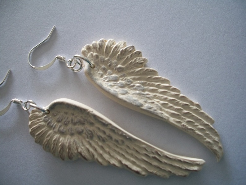 Puede incluir: Un par de pendientes de aro de plata con alas de &aacute;ngel de cer&aacute;mica blanca. Las alas tienen un dise&ntilde;o texturizado y plumoso.