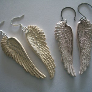 Puede incluir: Dos pares de pendientes de plata con forma de alas de &aacute;ngel. Las alas est&aacute;n hechas de un material blanco y tienen una superficie texturizada. Los pendientes cuelgan de ganchos de plata.