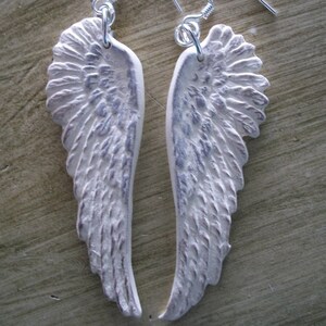 K&ouml;nnte beinhalten: Ein Paar wei&szlig;e Engelsfl&uuml;gel-Ohrringe mit silbernen Haken. Die Fl&uuml;gel sind texturiert und haben einen leicht abgenutzten Look.