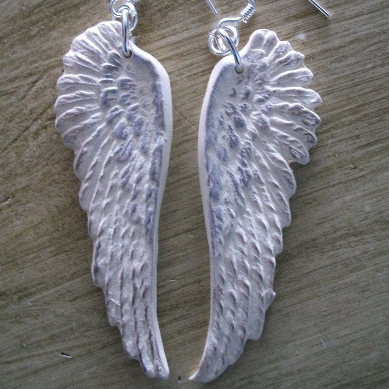 K&ouml;nnte beinhalten: Ein Paar wei&szlig;e Engelsfl&uuml;gel-Ohrringe mit silbernen Haken. Die Fl&uuml;gel sind texturiert und haben einen leicht abgenutzten Look.