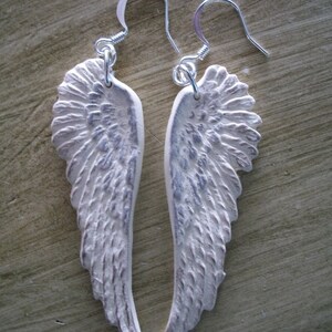 K&ouml;nnte beinhalten: Ein Paar silberne Ohrringe mit wei&szlig;en Engelsfl&uuml;geln. Die Fl&uuml;gel sind strukturiert und haben einen Used-Look.