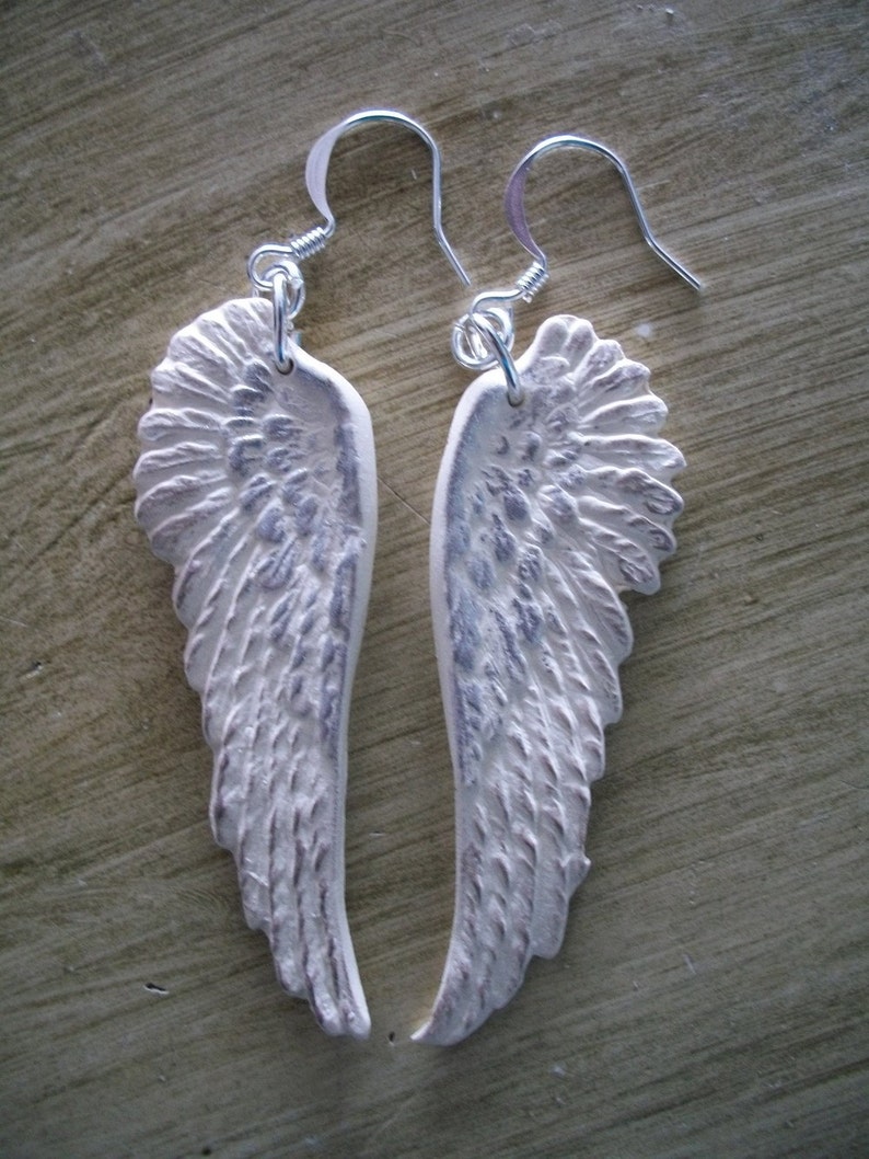 K&ouml;nnte beinhalten: Ein Paar silberne Ohrringe mit wei&szlig;en Engelsfl&uuml;geln. Die Fl&uuml;gel sind strukturiert und haben einen Used-Look.