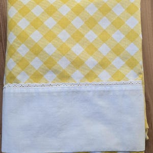 May include: A folded yellow and white gingham patterned sheet with a white border and lace trim. The sheet is made of a soft fabric and is likely for a bed. The overall theme is home textiles.