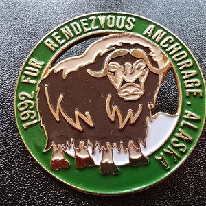 May include: A circular green and gold metal pin with the text "1992 FUR RENDEZVOUS ANCHORAGE ALASKA". The pin features a brown and gold musk ox illustration in the center.
