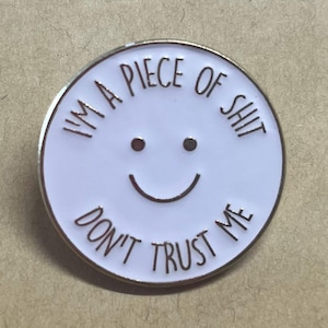 May include: A circular white enamel pin featuring a simple black smiley face in the centre. The text "I'M A PIECE OF SHIT" is printed in black along the top curve, and the text "DON'T TRUST ME" is printed in black along the bottom curve.