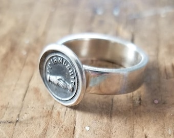Può includere: Un anello d'argento con un sigillo rotondo inciso che presenta una mano e la parola "Friendship".