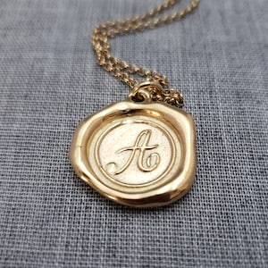 Può includere: Una collana con catena dorata con un ciondolo dorato a forma di sigillo di cera con la lettera "A" in rilievo.