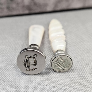 Puede incluir: Dos sellos de lacre plateados antiguos con mangos blancos. Un sello tiene un diseño de letra "E" adornada, el otro un diseño de escudo. Los sellos se muestran sobre una superficie gris.