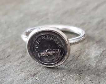 May include: A silver ring with a round, engraved seal that reads "Friendship". The seal features a design of two hands clasped together.
